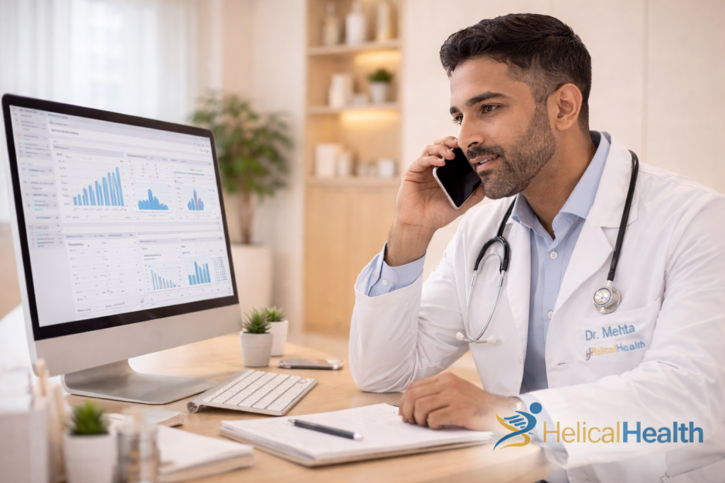 A male doctor wearing a white coat and stethoscope sits at a desk in a bright, modern office, speaking on a smartphone while looking toward a large desktop monitor. The screen displays charts, graphs, and data dashboards. A notepad, pen, keyboard, and small potted plant rest on the desk. His coat reads “Dr. Mehta” with the HelicalHealth logo, and the “HelicalHealth” logo also appears in the bottom right corner.